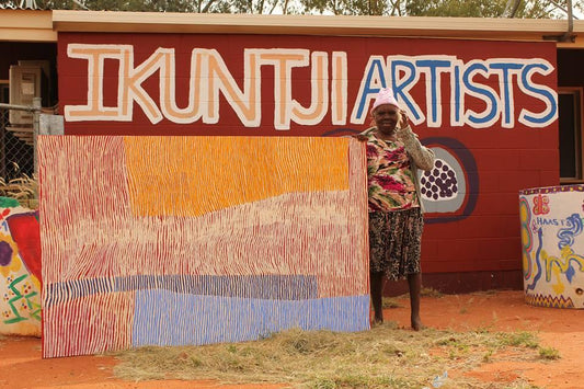 Artist holding a large Indigenous artwork painting in front of a wall with 'Ikuntji Artists' painted on the side